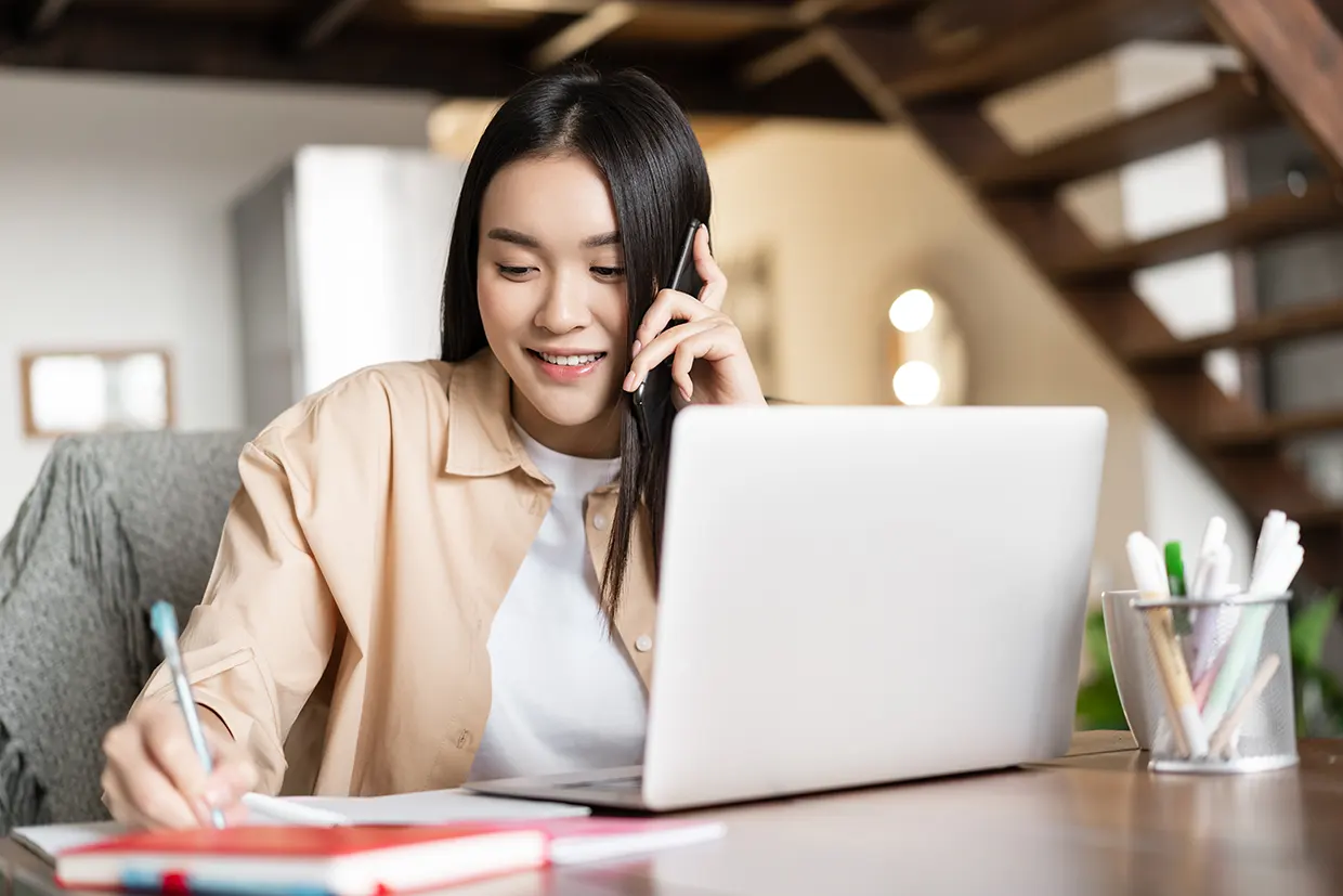 businesswoman at laptop smiling and taking notes on phone to digital agency